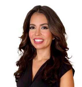 A woman with long, wavy dark hair and a bright smile is wearing a black sleeveless top. She stands against a plain white background.